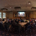 A fisheye lens view from the back of a room of a person speaking in the front while dozens of seated people are listening; an image is displayed on the screen behind the speaker